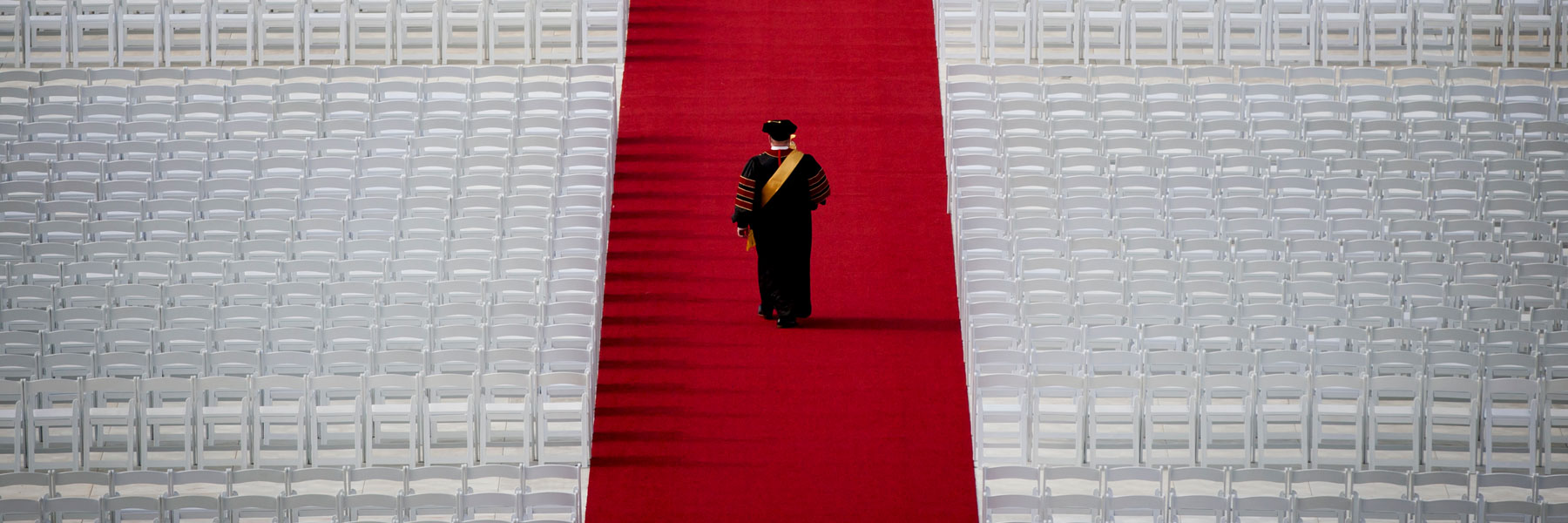 A dramatic aerial photograph of Indiana University's Grand Marshal walking down a red carpet. He is flanked by rows of  empty white seats. 