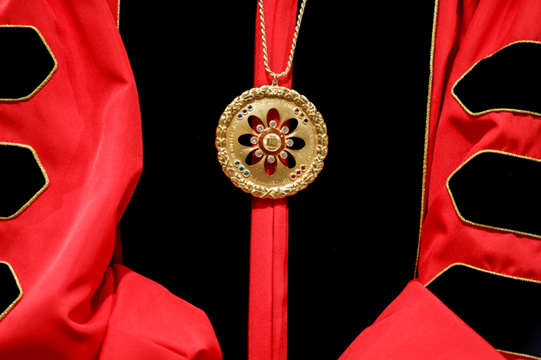 A close-up of the jewel of Indiana University worn by Dr. Pamela Whitten, 19th president of Indiana University. The jewel is a golden medallion wreathed with bound laurel motifs and stamped with gold lettering. At its center is the university seal, surrounded by diamonds, with colored gems at the compass points. 