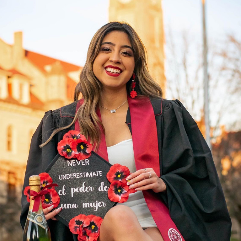A young woman with wavy highlighted hair and red lipstick holds a brightly colored mortarboard decorated with flowers. It reads "never underestimate el poder de una mujer" (translated, from Spanish, the power of a woman).