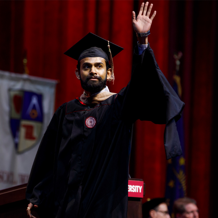 A male graduate student from the Kelley School of Business, wearing master's graduation attire, waves to the audience. The unique style of his gown's sleeve is on display. 